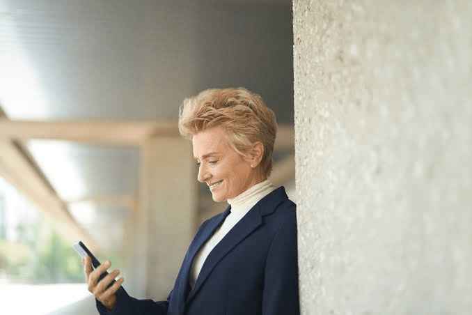 Middle-aged woman with short blonde hair wearing a white turtleneck and navy blue blazer, smiling while looking at her smartphone and leaning against a concrete pillar outdoors.